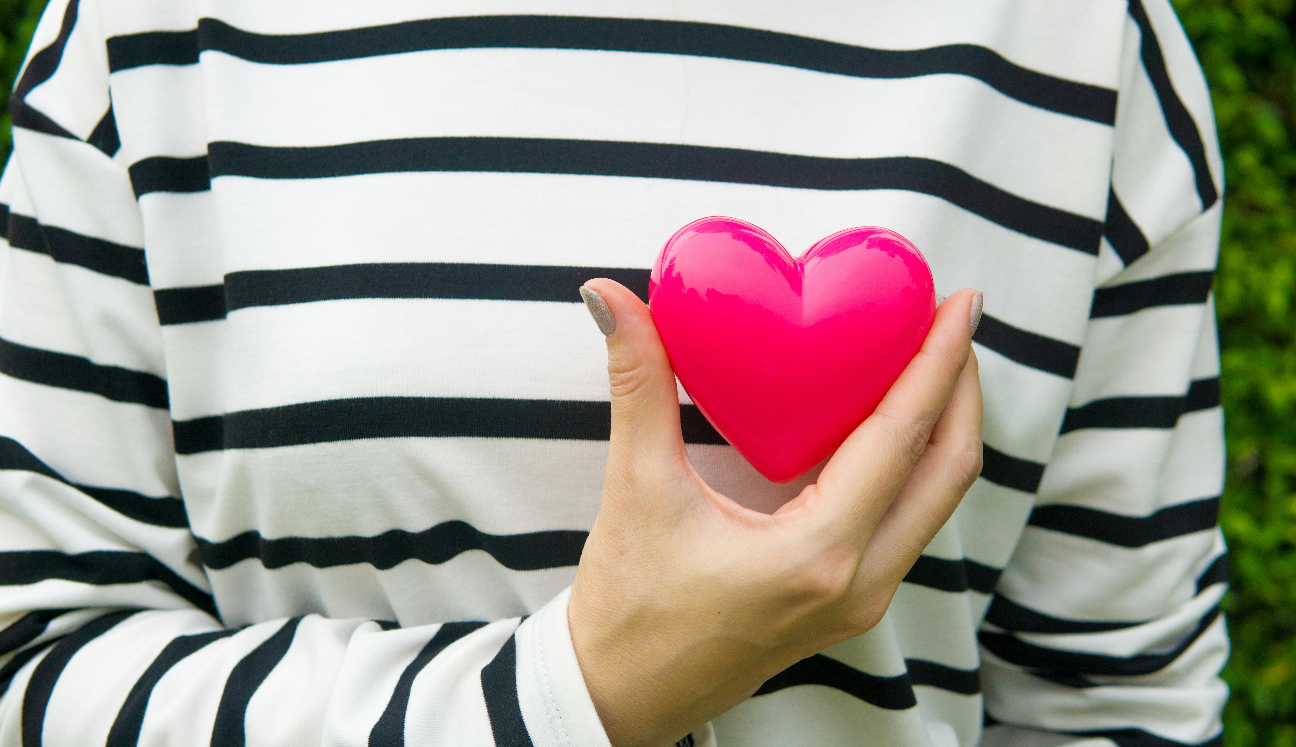 a person in a white sweater with thin black stripes holds a pink Valentine's day style heart in front of their chest